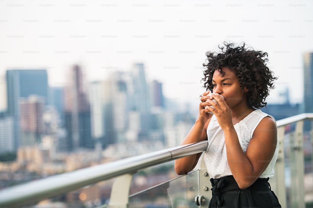 Ein Porträt einer schwarzen Frau, die auf einer Terrasse steht und Kaffee trinkt. Speicherplatz kopieren.