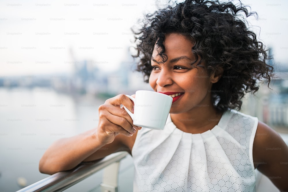 A portrait of a black woman standing on a terrace, drinking coffee.