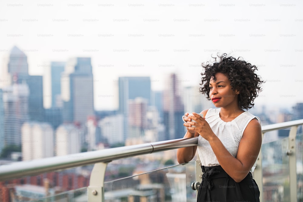 Portrait d’une femme noire debout sur une terrasse, tenant une tasse de café. Espace de copie.