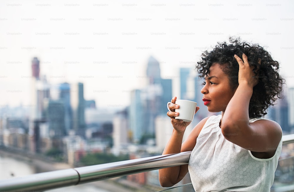 Un ritratto ravvicinato di una donna nera in piedi su una terrazza, bevendo caffè. Copia spazio.