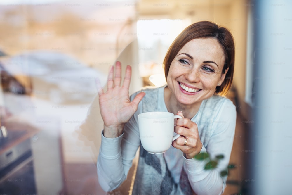 Una giovane donna con una tazza di caffè che guarda fuori da una finestra, salutando il marito al mattino. Girato attraverso il vetro.