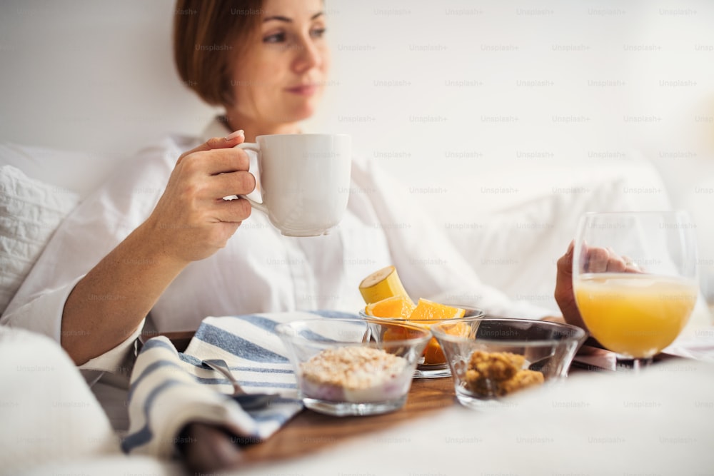 Eine junge Frau, die morgens mit Kaffee und Frühstück drinnen in einem Schlafzimmer im Bett liegt.