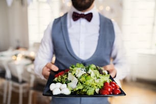 A midsection of man standing indoors in a room set for a party, holding a tray with vegetables.