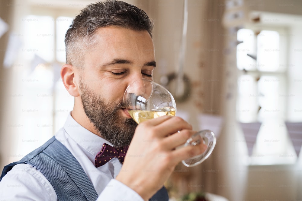 Un ritratto di uomo maturo hipster in casa in una stanza allestita per una festa, bevendo vino bianco.