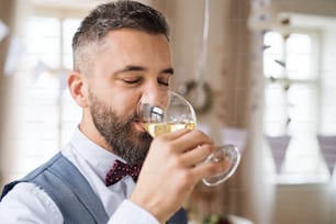 A portrait of hipster mature man indoors in a room set for a party, drinking white wine.