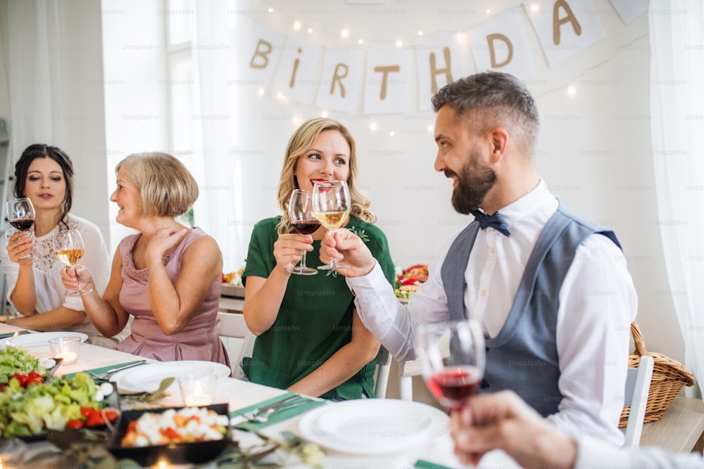 Une grande famille assise à une table lors d’une fête d’anniversaire à l’intérieur, faisant claquer des verres avec du vin.