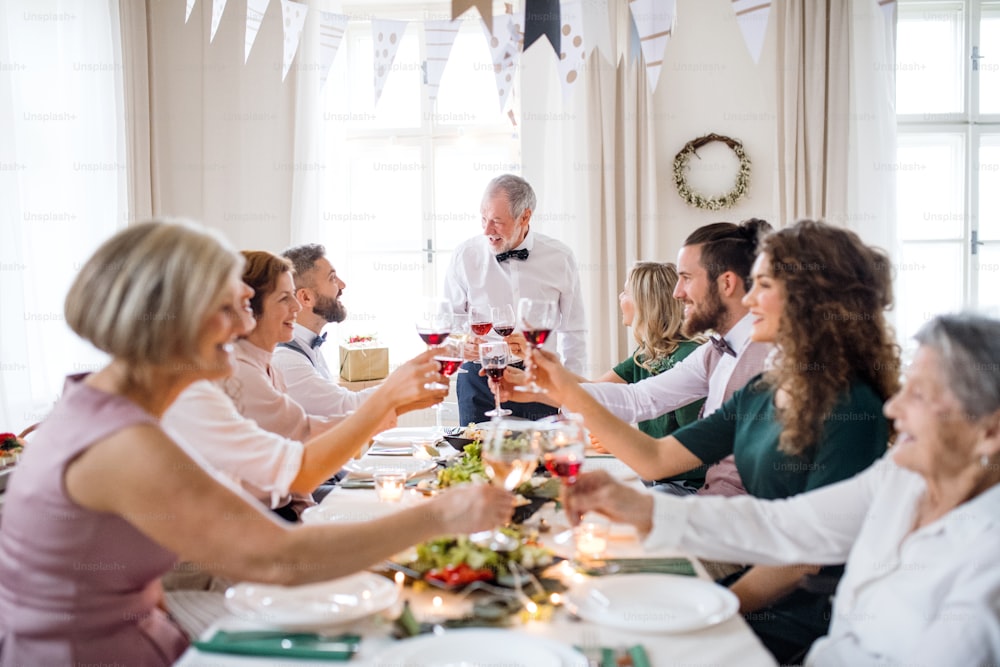 Une grande famille assise à une table lors d’une fête d’anniversaire à l’intérieur, faisant claquer des verres avec du vin rouge.