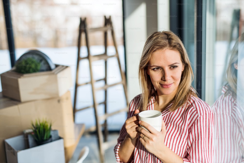Una giovane donna felice che si trasferisce in una nuova casa, appoggiata a una finestra e bevendo caffè.