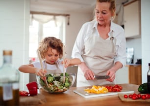 A portrait of small girl with grandmother in a kitchen at home, preparing vegetable salad.