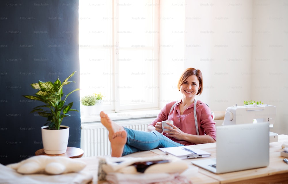 Junge kreative Frau mit Kaffee in einem Studio, die sich mit den Füßen auf dem Schreibtisch ausruht. Ein Startup eines kleinen Schneidergeschäfts.