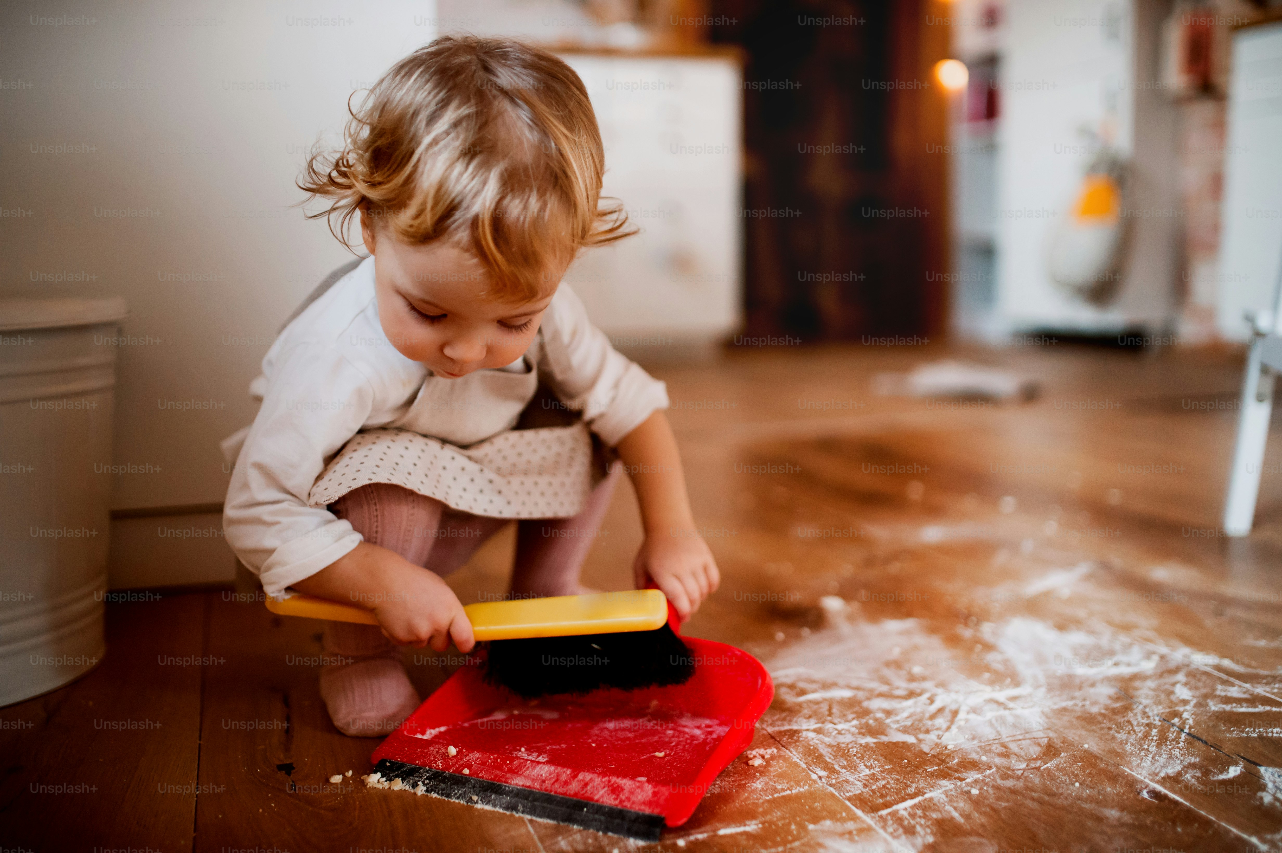 A small toddler girl with brush and dustpan sweeping messy floor in the kitchen at home