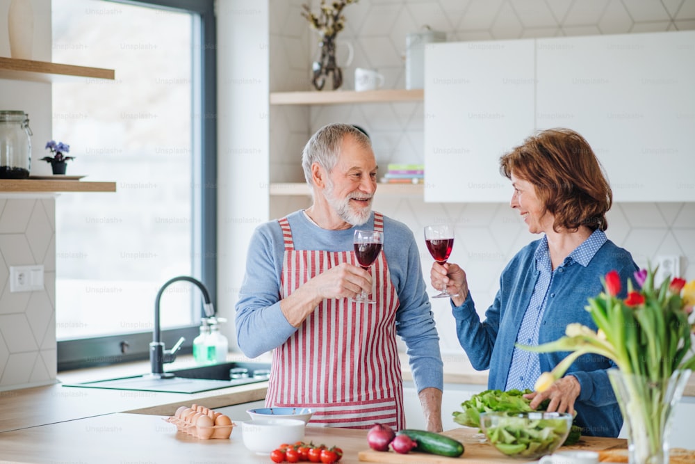 Um retrato de um casal idoso apaixonado dentro de casa, piscando taças com vinho ao cozinhar.