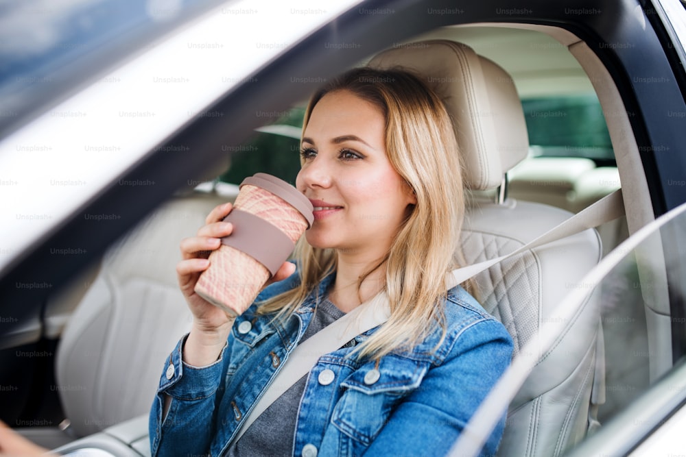 車に座って紙コップからコーヒーを飲む若い女性ドライバー。