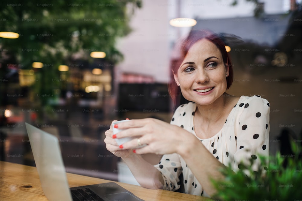 Ein Porträt einer Frau mit Kaffee, die am Tisch in einem Café sitzt und einen Laptop benutzt. Durch Glas geschossen.