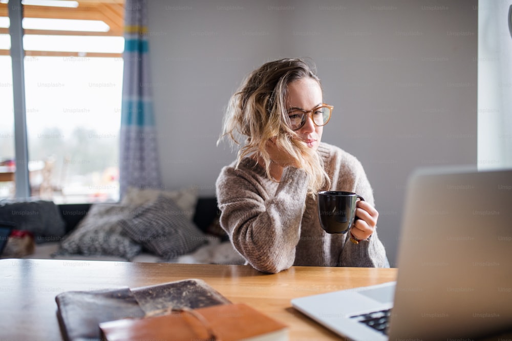 Portrait d’une jeune femme avec un ordinateur portable et du café travaillant à l’intérieur à la maison.