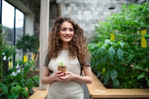 Portrait of woman gardener standing in greenhouse, holding small plant.