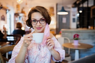 Portrait of young woman with face mask and coffee indoors in cafe, looking at camera.