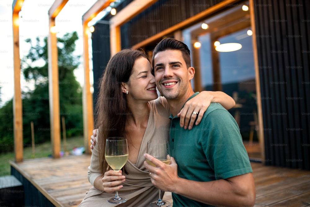 Giovane coppia con vino seduto all'aperto, fine settimana in casa contenitore in campagna.