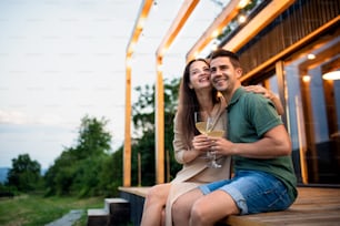 Jeune couple avec vin assis à l’extérieur, week-end dans une maison de conteneur à la campagne.