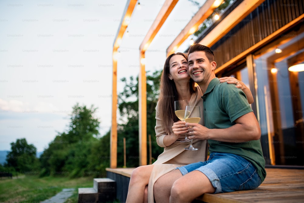 Giovane coppia con vino seduto all'aperto, fine settimana in casa contenitore in campagna.
