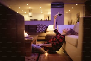 Uma jovem feliz sentada no chão e bebendo vinho à noite em casa sozinha.