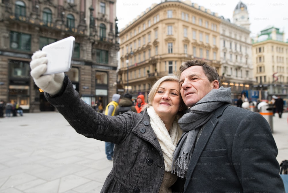 Bella coppia anziana in vestiti invernali durante una passeggiata nel centro storico della città di Vienna, Austria. Donna che scatta selfie di loro con lo smartphone.