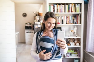 Beautiful young mother with her newborn baby son in sling taking selfie with smart phone