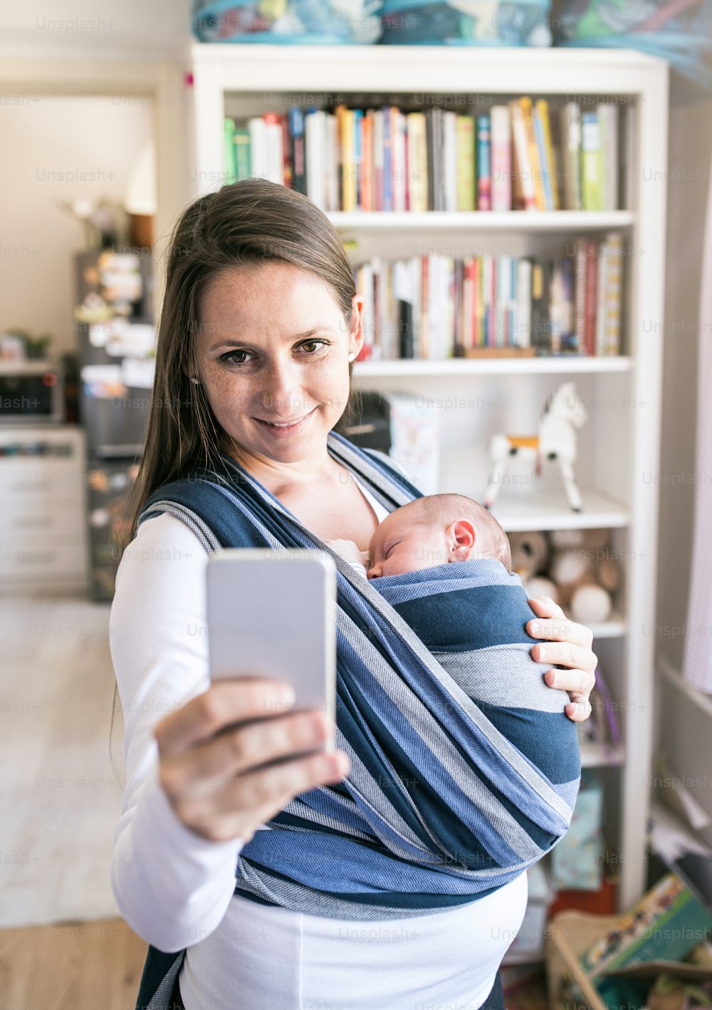 Beautiful young mother with her newborn baby son in sling taking selfie with smart phone