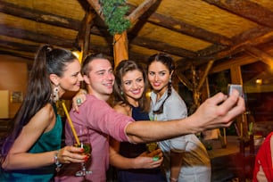 Young beautiful people with cocktails in bar or club taking selfie, having fun