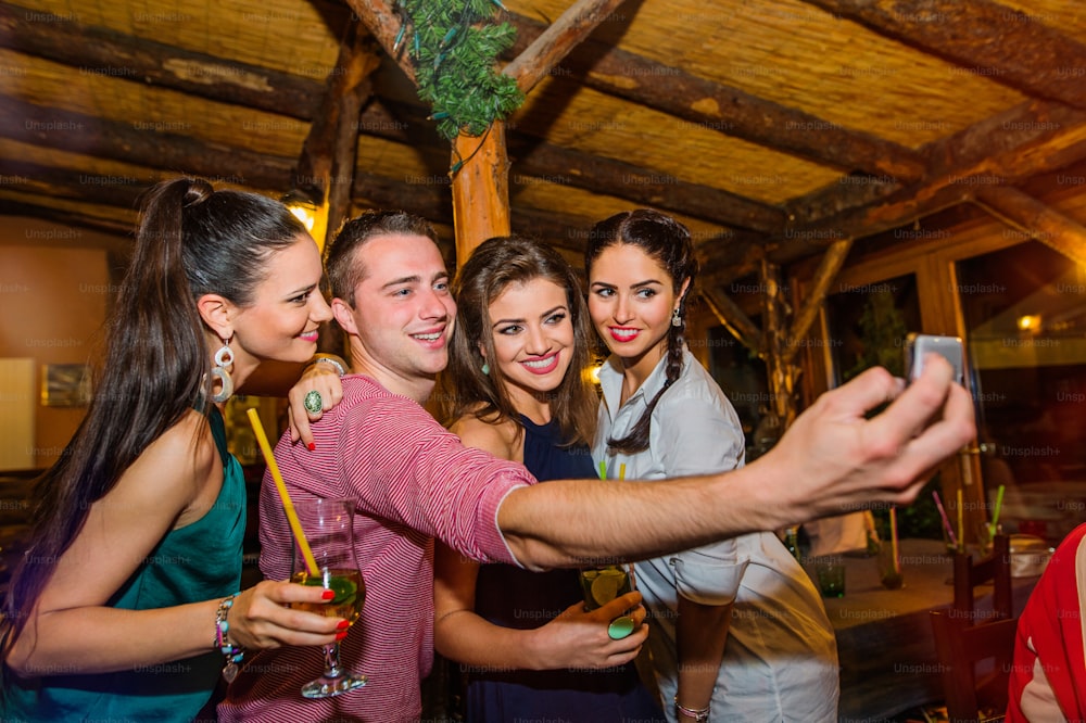 Young beautiful people with cocktails in bar or club taking selfie, having fun