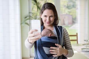 Beautiful young mother with her newborn baby son in sling at home, holding smart phone, taking selfie