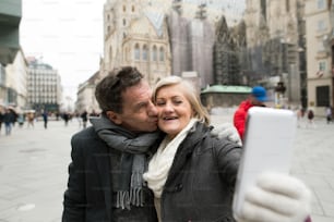 Schönes älteres Paar bei einem Spaziergang im historischen Zentrum der Stadt Wien, Österreich. Frau macht Selfie von ihnen mit Smartphone. Ehemann gibt seiner Frau einen Kuss. Winter.