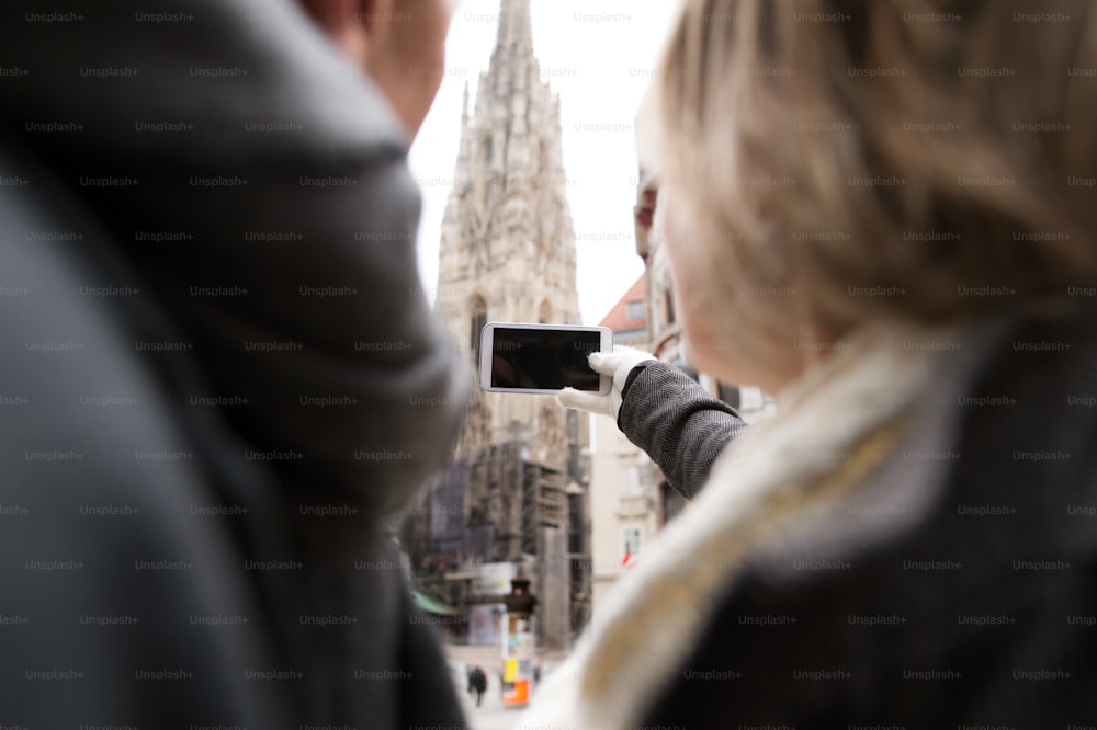 Nicht wiederzuerkennendes älteres Ehepaar bei einem Spaziergang im historischen Zentrum der Stadt Wien, Österreich. Frau macht Selfie von ihnen mit Smartphone. Winter.