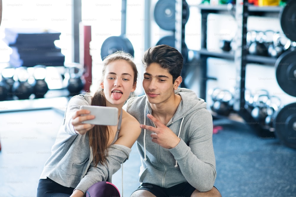 Beau jeune couple en forme dans une salle de gym moderne avec téléphone intelligent, prenant selfie.