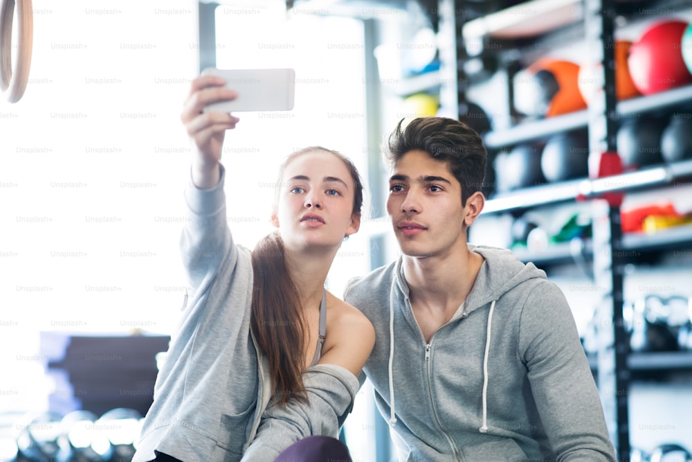 Lindo casal jovem em forma no ginásio moderno ginásio com telefone inteligente, tirando selfie.