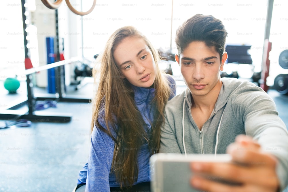 Beau jeune couple en forme dans une salle de sport moderne avec téléphone intelligent, prenant selfie.