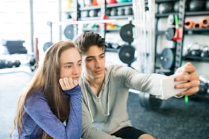 Beautiful young fit couple in modern  gym with smart phone, taking selfie.