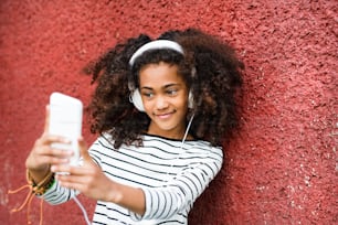 Beautiful african american girl with curly hair, holding smart phone, wearing headphones, listening music and taking selfie.