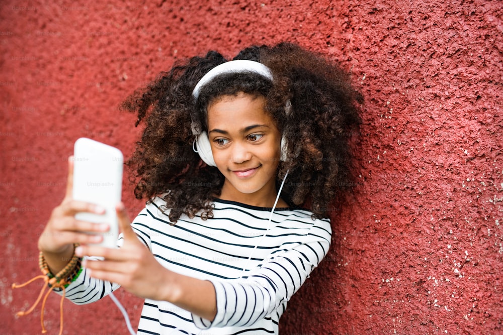 Beautiful african american girl with curly hair, holding smart phone, wearing headphones, listening music and taking selfie.