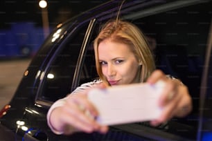Hermosa joven en una ciudad en su automóvil moderno por la noche, sosteniendo un teléfono inteligente, tomándose selfie.