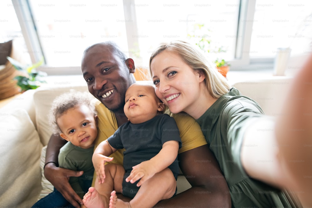Beautiful young interracial family at home holding their cute son and daughter in the arms, taking selfie.