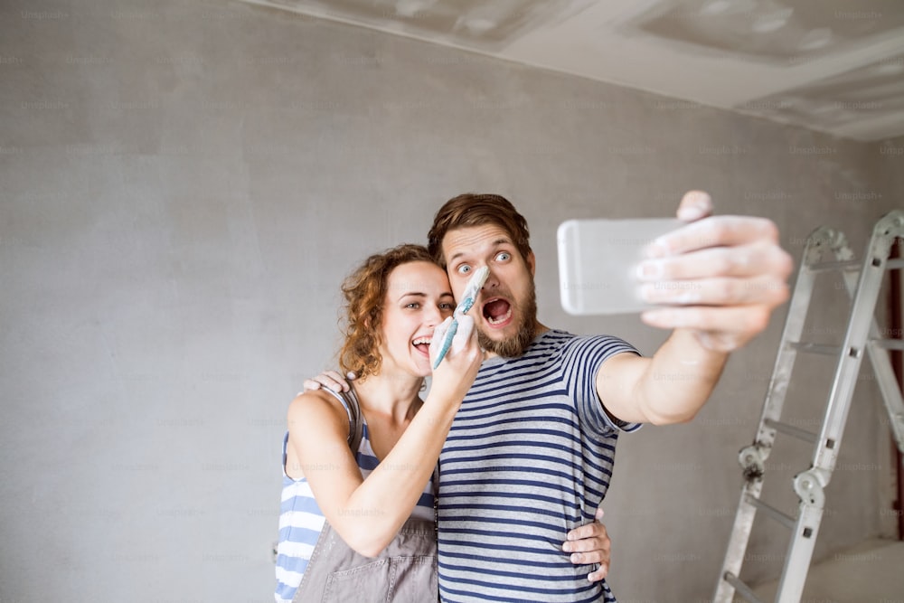 사랑에 빠진 아름다운 젊은 부부는 새 집의 벽을 칠하고, 스마트폰으로 셀카를 찍는다. 집 화장 및 개조 개념입니다.