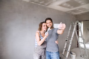 Beautiful young couple in love having fun, painting walls of their new house, taking selfie with smart phone. Home makeover and renovation concept.