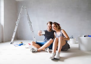 사랑에 빠진 아름다운 젊은 부부는 새 집의 벽을 칠하고, 스마트폰으로 셀카를 찍는다. 집 화장 및 개조 개념입니다.