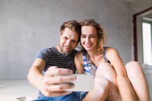 Lindo jovem casal apaixonado se divertindo, pintando paredes de sua nova casa, tirando selfie com smartphone. Conceito de reforma e renovação da casa.