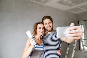 Schönes junges verliebtes Paar, das Spaß hat, die Wände ihres neuen Hauses streicht und Selfies mit dem Smartphone macht. Umbau- und Renovierungskonzept.