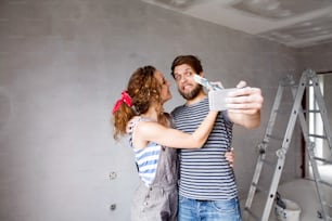 Beau jeune couple amoureux s’amusant, peignant les murs de leur nouvelle maison, prenant selfie avec un téléphone intelligent, faisant drôle de visage. Concept de rénovation et de rénovation de la maison.
