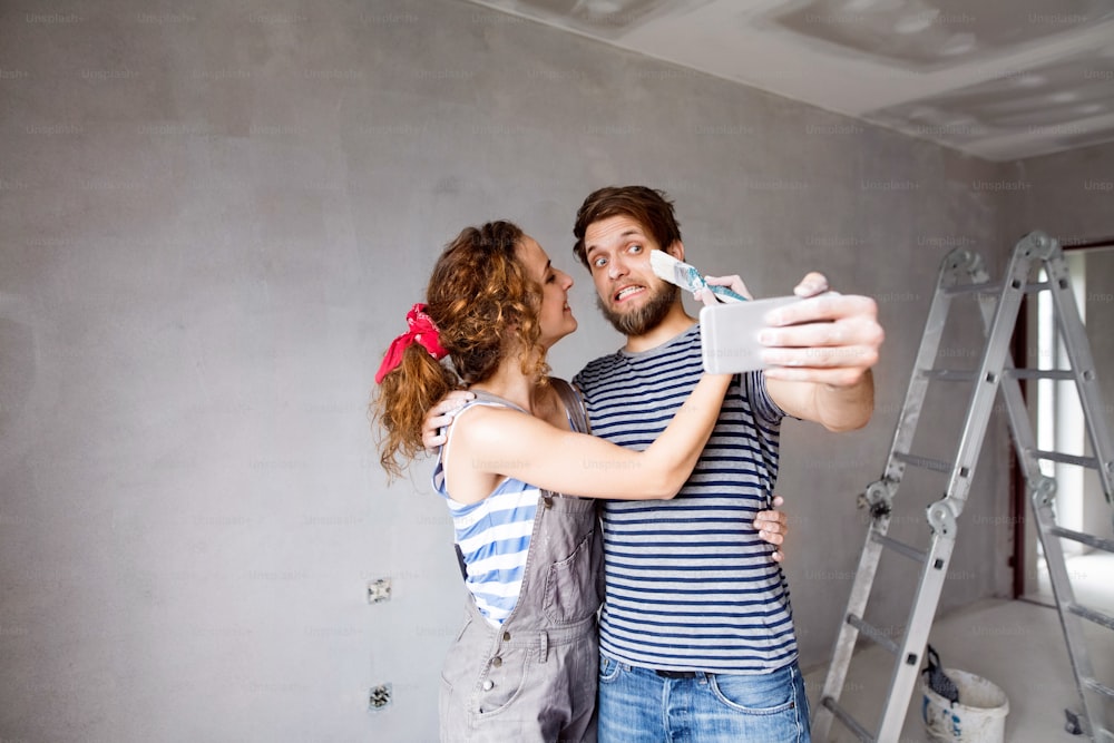 Beautiful young couple in love having fun, painting walls of their new house, taking selfie with smart phone, making funny face. Home makeover and renovation concept.