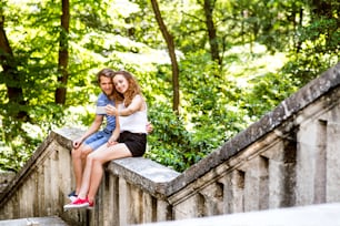 Hermosa pareja joven en la ciudad sentada en una pared de concreto, tomándose selfie con un teléfono inteligente. Soleado día de primavera.