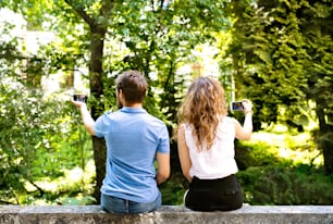 Hermosa pareja joven con teléfonos inteligentes sentados en una pared de concreto, tomando selfies. Soleado día de primavera. Vista trasera.